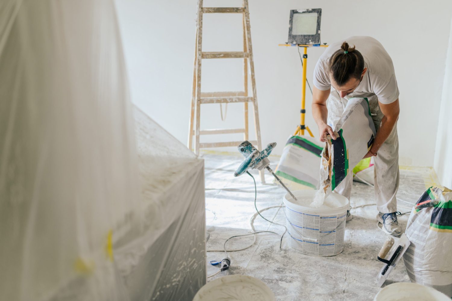 A builder is standing in a house in renovation process and making plastering materials for skim coating walls. A constructor is adding plaster into a bucket and making plaster paste for plastering.